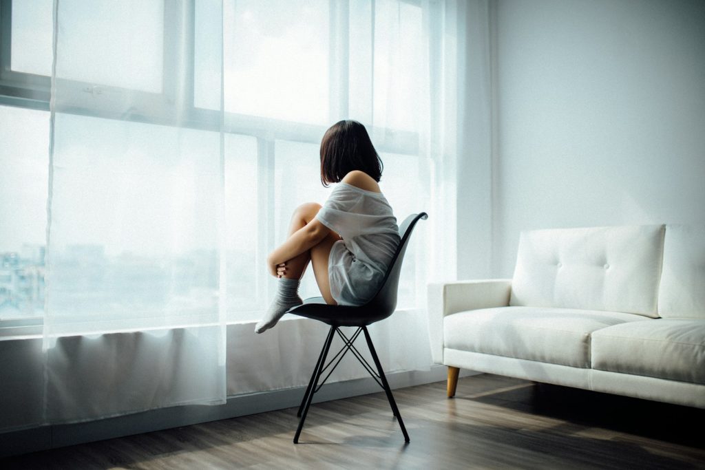 woman sitting on black chair in front of glass-panel window with white curtains experiencing the Emotional Impact of Cancer Diagnosis